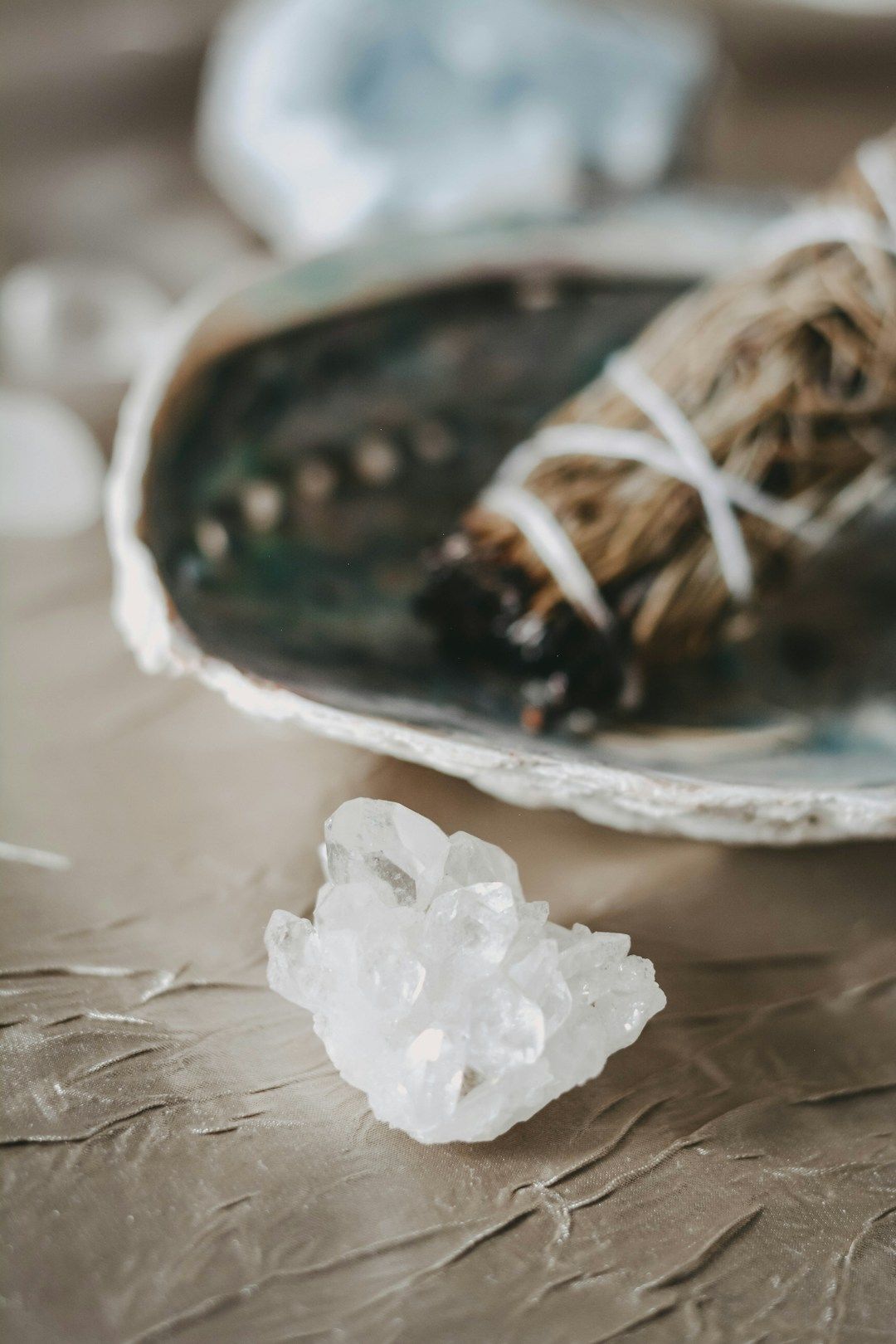 A white crystal is shown on a silver surface ink front of a blurred, big shell that holds a smudge stick. 