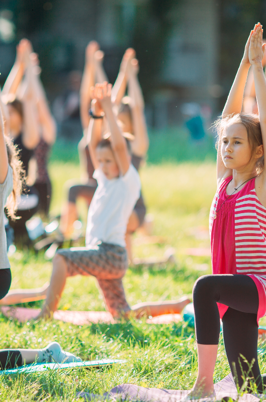 A group of children is shown on a lawn, all in low Lunge with the right leg bent at 90 degrees in front of them, both arms raised straight overhead, palms touching together gently.