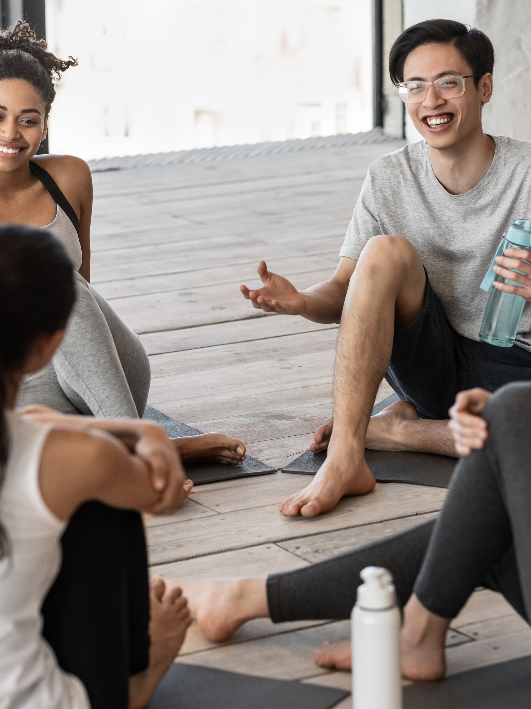 A group of yoga students sit in a circle on their mats, holding a lively discussion. A smiling woman is listening as a man gestures enthusiastically.
