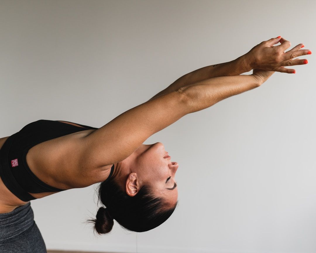 A close-up of a woman in a standing backbend with her arms extended overhead and behind her. Her dark hair is pulled back into a low bun and her eyes are closed in focus. She is wearing a black athletic top and gray leggings against a light gray background.