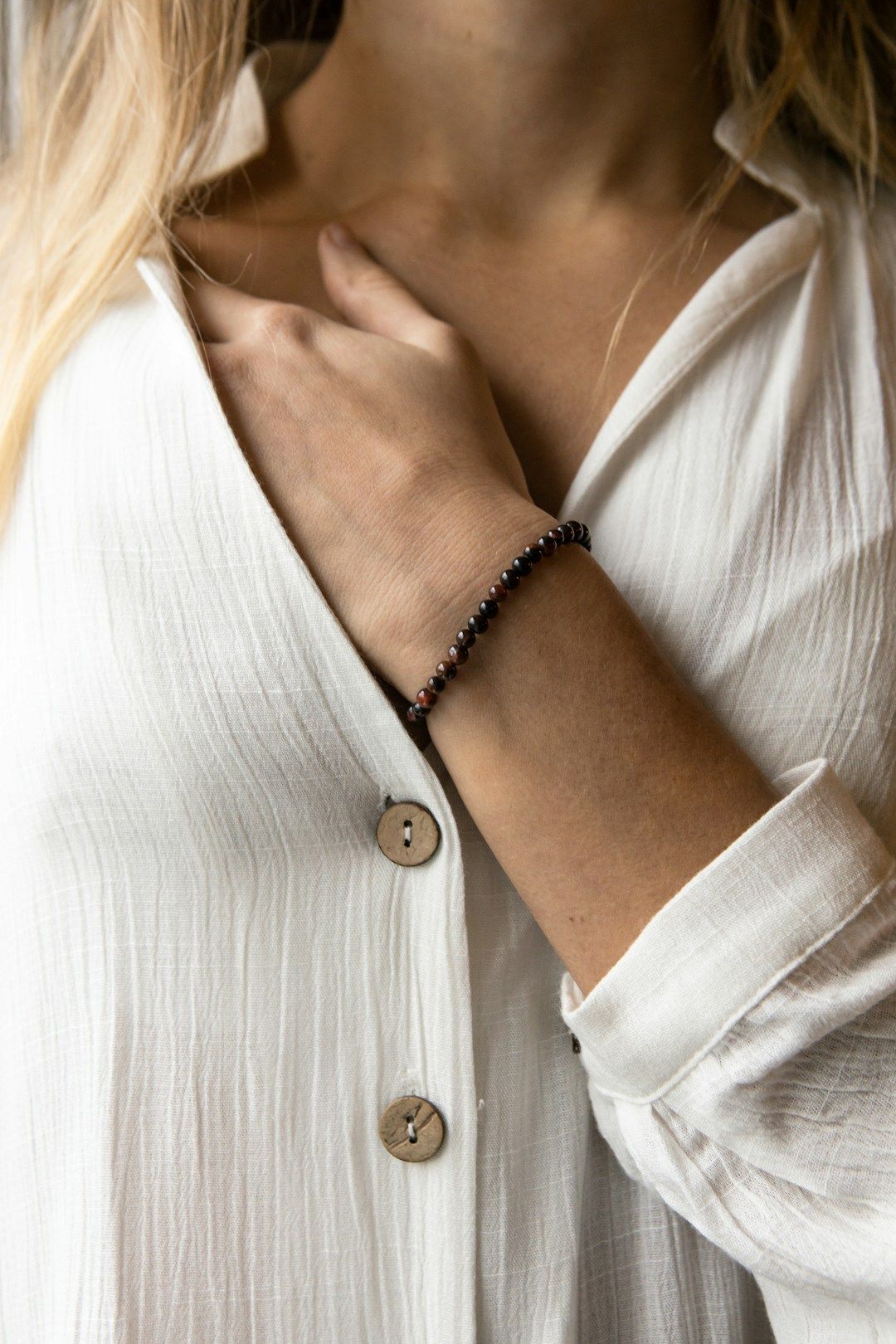 Someone will long blong hair is shown resting their left hand on chest level und a white cotton tunic with natural brown buttons.