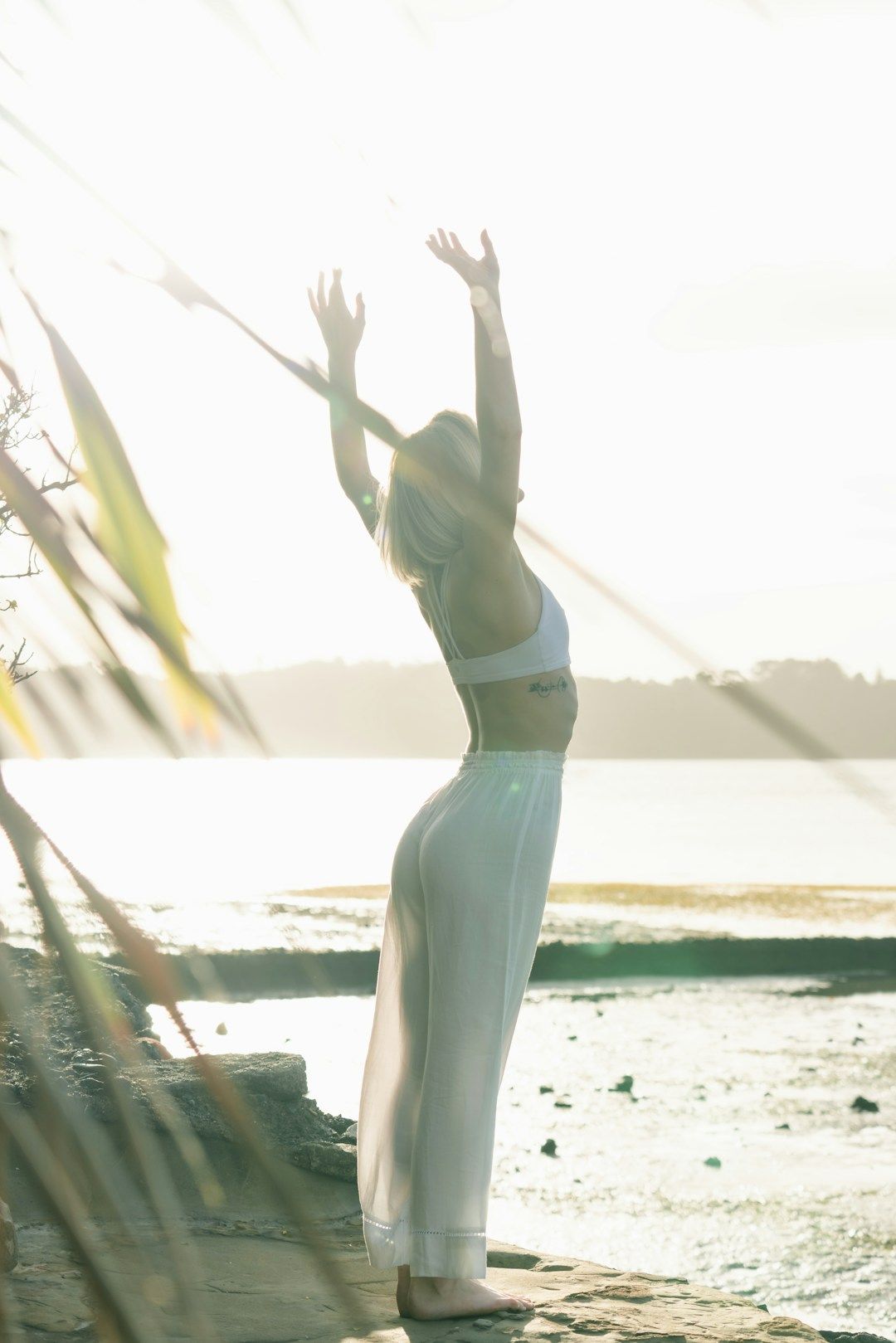 A woman stands on a beach, with her arms raised into the air. She is wearing white linen pants and a white athletic crop top. She is softly lit by golden light.