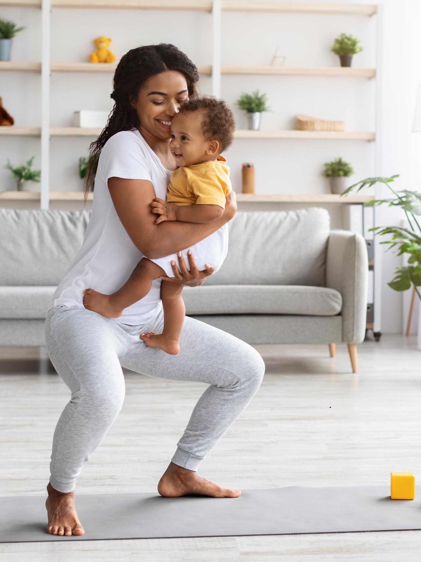 A woman is pictured holding her baby while in a high squat on a yoga mat. She is wearing gray sweatpants and a white shirt with her baby in her arms. She is smiling and has her curly black hair casually framing her face. 