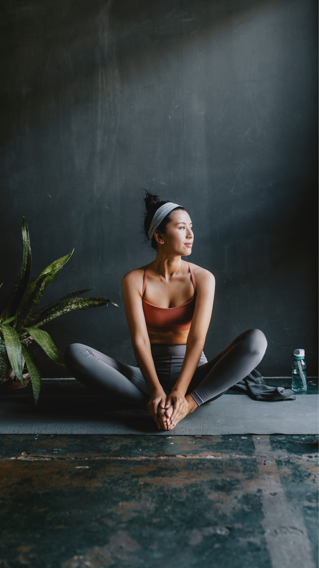 A woman sits on the ground in front of her laptop with her legs crossed. She has her left hand on the opposite knee in a stretch. She has curly hair and sits in front of a green plant. There are candles lit around her.