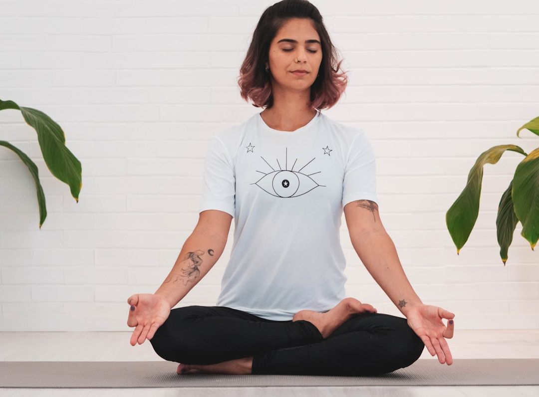 A yogi is sitting in Lotus Pose with their hands resting on their knees, palms facing upwards and thumbs and index fingers pressing together gently. They are wearing a white shirt with the outline of an eye, black leggings, and have dark hair with pink ends. Their eyes are closed and they’re shown against a white brick wall with plants on either side. 