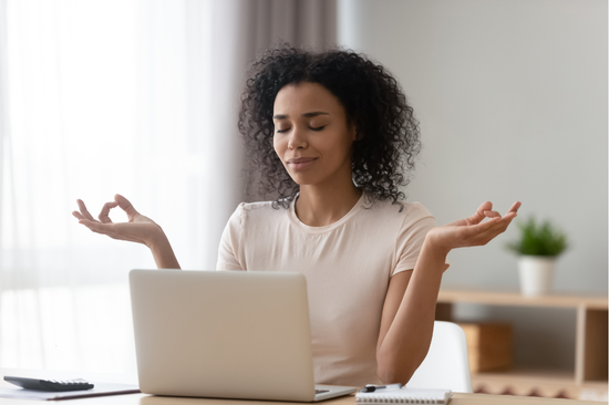 A woman with curly hair sits at a desk in front of a laptop, eyes closed and hands raised in a relaxed meditation pose, with a notebook and pen nearby in a softly lit room.