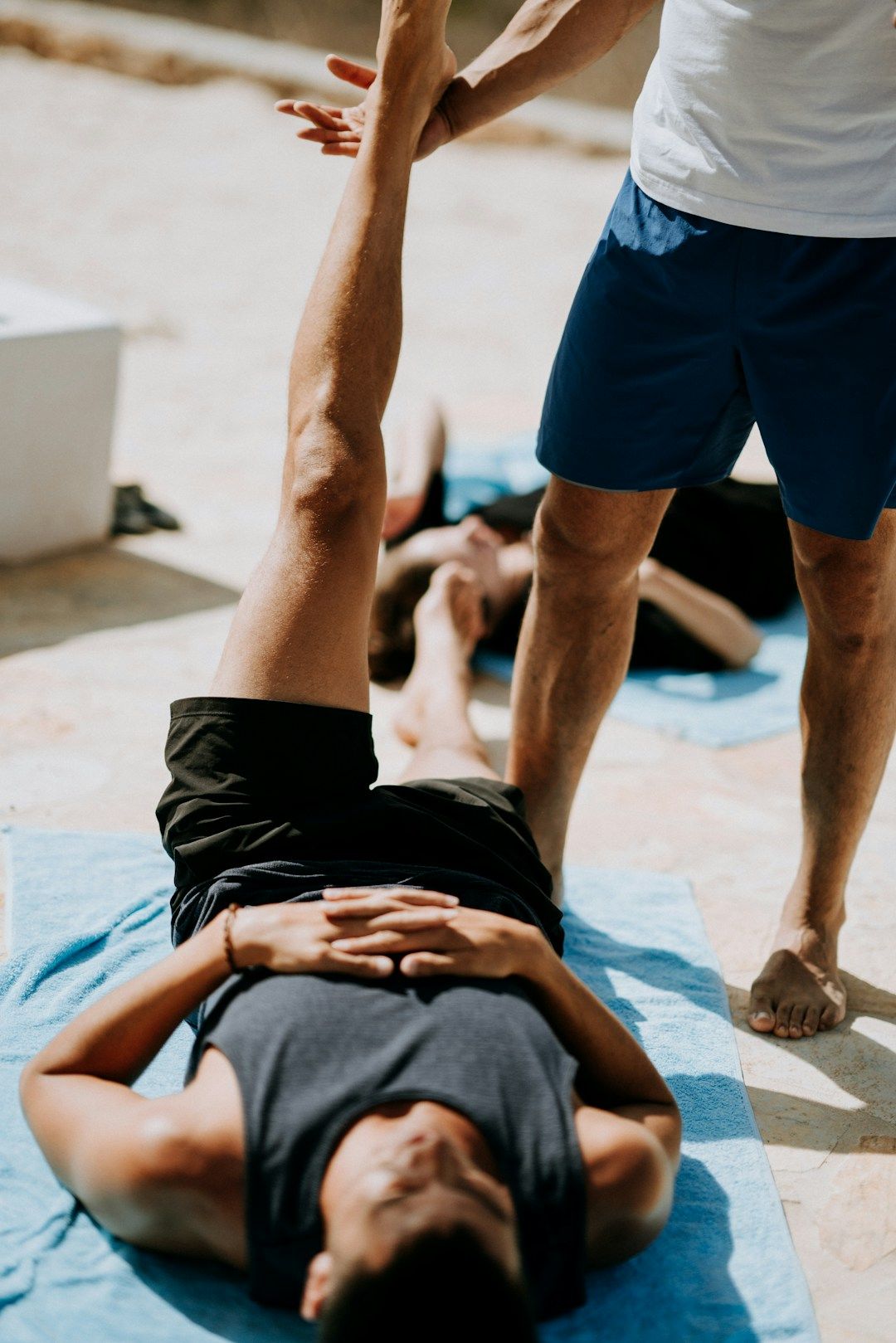 * A man is resting on a towel on the ground, the left leg raised straight upward. A second man is offering assistance with one hand at the first man’s foot.