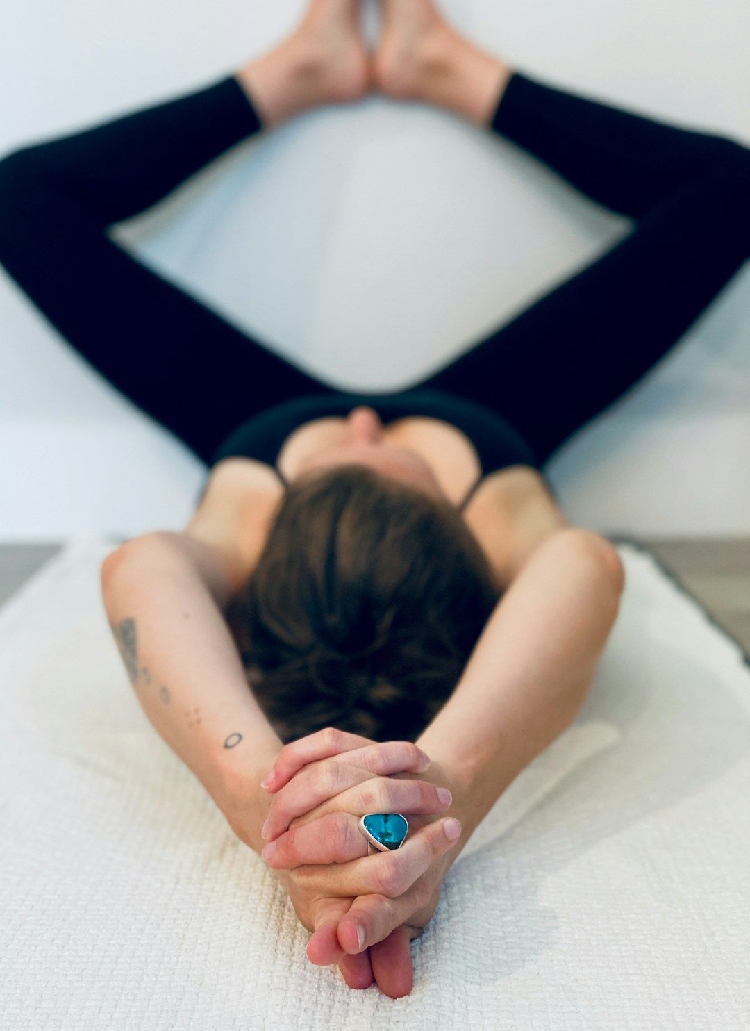 A yogi is shown resting on their back, their legs raised at a white wall and the soles of their feet touching in an elevated Butterfly pose. They are wearing all black and have their hands stretched backward over their head, fingers clasped together. 