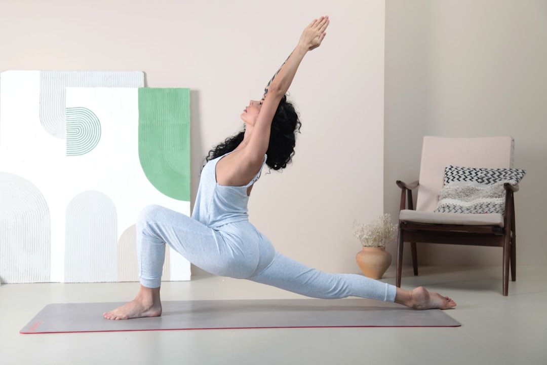 A yogi is shown on a light grey mat against a beige wall in Low Lunge Pose, the arms stretched above and beyond their head, palms touching. They are wearing a grey sports set and are shown in a room with two abstract art pieces and a dark wooden chair with light beige sitting pillows. 