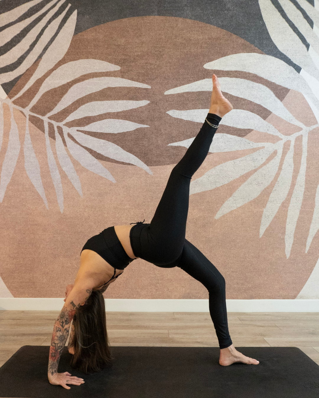 A yogi dressed in all black with a sleeve tatto on their right arm is shown in bridge pose, with their right leg lifted up. They are pictured against a leavy print wallpaper in earthen colors.