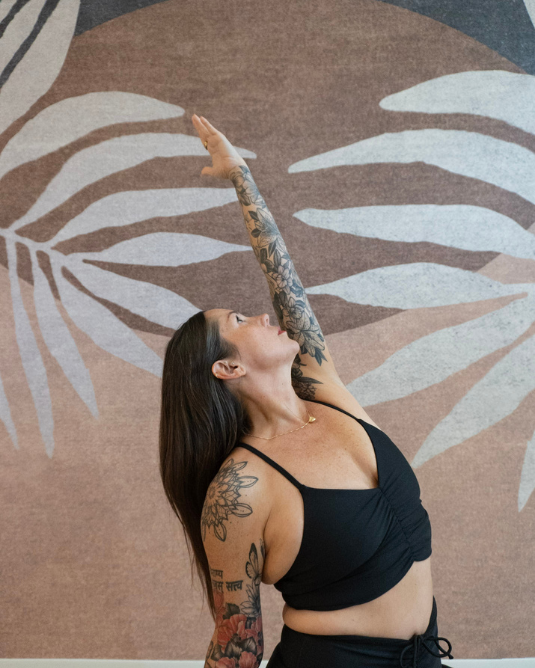 A woman practicing yoga in a studio, performing a yoga asana. She has her arm raised into the air, with her gaze directed toward her hand. She has long hair, tattoos on her arms, and is wearing a black sports bra and leggings. The background features soft pastel colors.