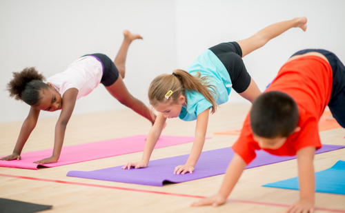 Three children on colourful mats in a studio are shown in Downward dog, two lifting their left leg behind and above them. 
