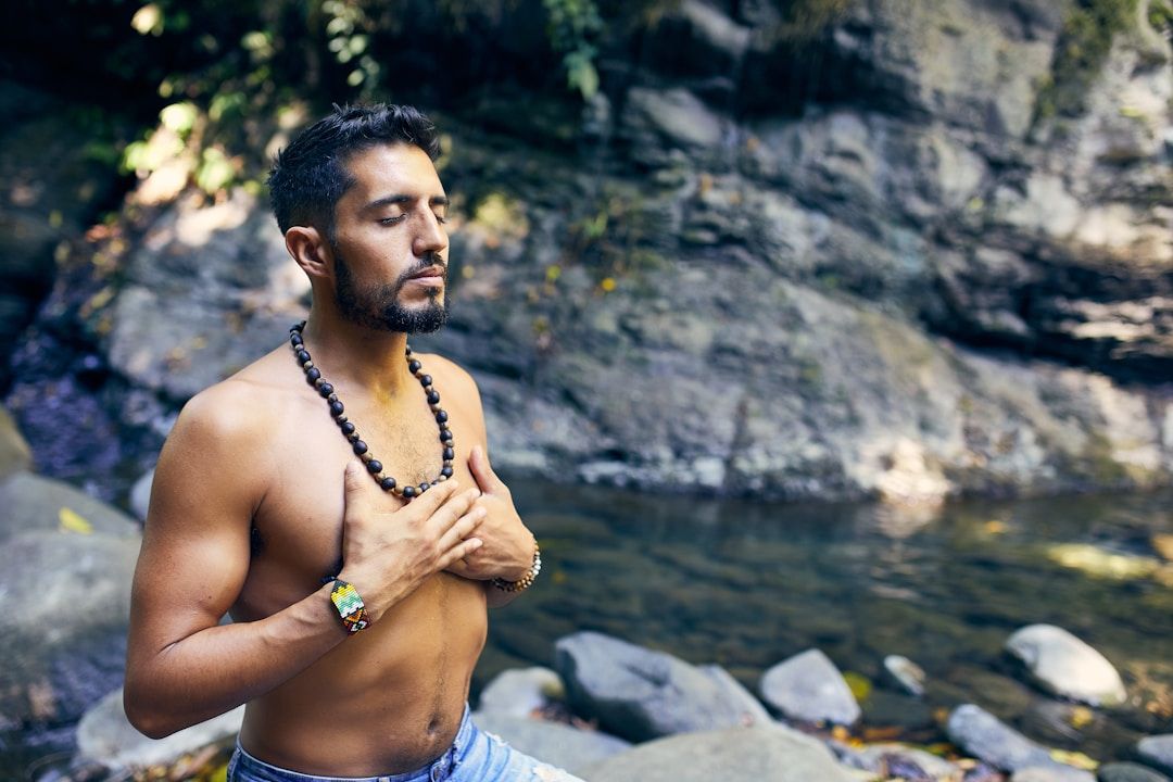 A man is shown in front of a creek, hands folded over the heart and eyes closed. He's topless and ha a beaded necklace.