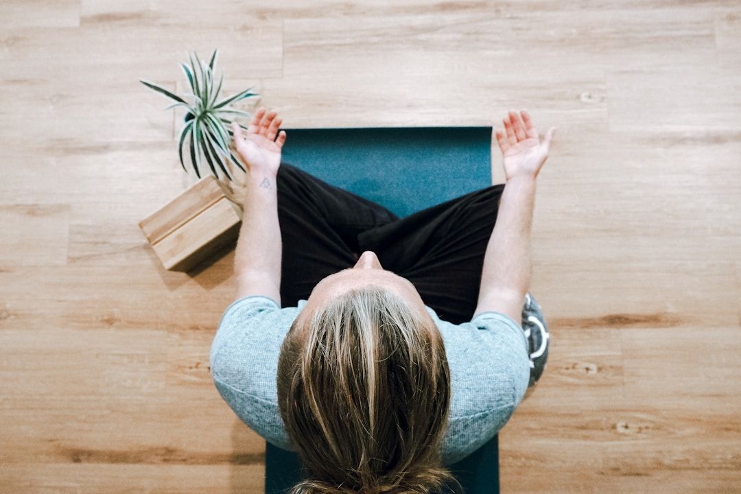 A yogi is sitting with legs crossed in front of them on their yoga mat on the ground. The camera is facing them from above. Their hands are resting on their knees, palms facing upward. To their right are two wooden yoga blocks and a plant.