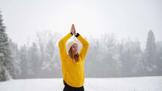 A woman stands outdoors in a snowy landscape, wearing a bright yellow sweater and a white knit hat. She holds her hands together overhead in a yoga pose, with snow-covered trees and a soft gray winter sky in the background. The scene conveys calm and mindfulness in a cold, peaceful setting.