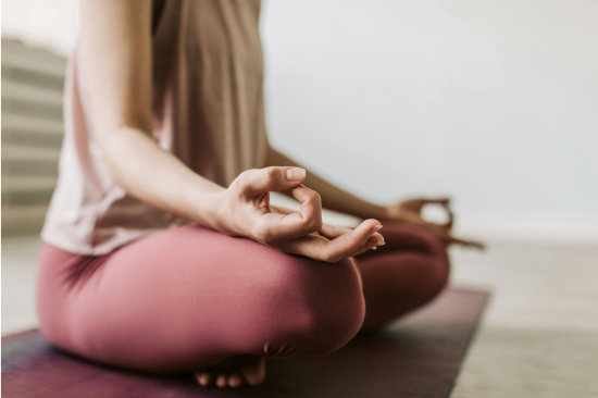 A woman sit cross-legged on a maroon yoga mat. Her hands are in a mudra position.