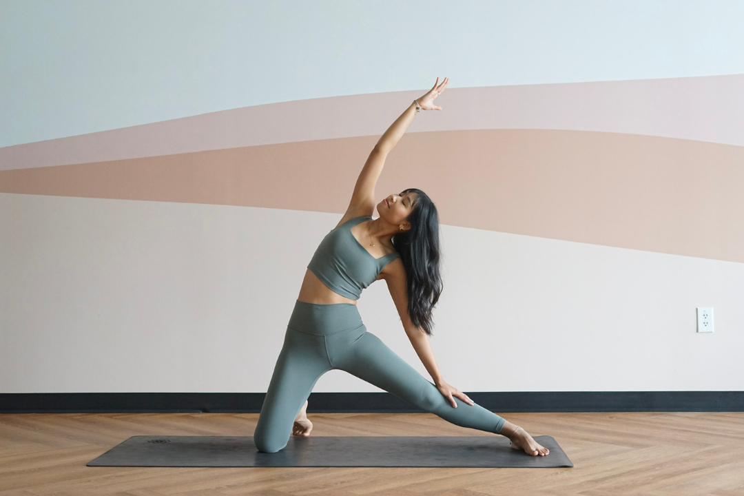 A yogi dressed in a pastel petroleum sports set is shown in Gate Pose, kneeling on their right knee on a black mat on wooden floors. Their left leg is stretched sideways at a 90 degree angle, their left hand resting on the calf, their right hand lifted high over head, bending the torso slightly to the left. The wall behind them is white and has streaks of pastel pink, orange, and yellow.
