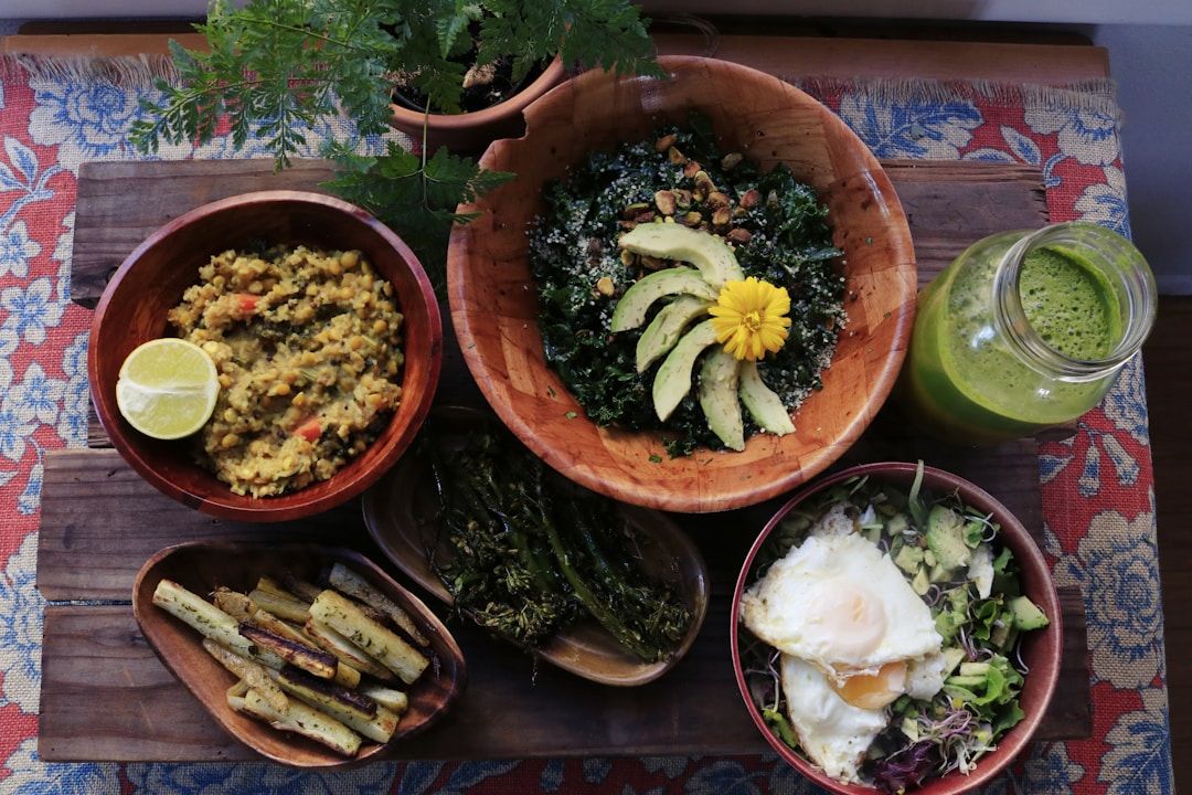 A wooden board on a Colourful table cloth, holding multiple wooden and ceramic bowls with healthy greens, quinoa, eggs, and more. There’s also. Mason jar with. Green smoothie and a decorative plant.