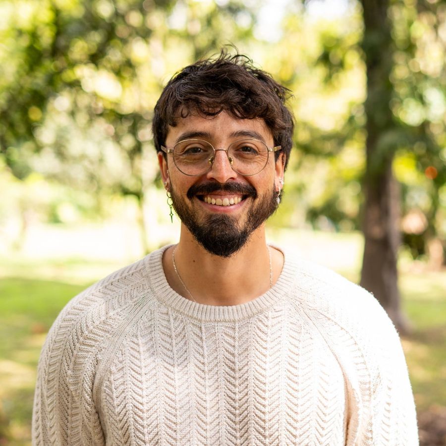 Retrato de Andrés Esteban Cárdenas, sonriente, con un suéter claro, en un parque arbolado