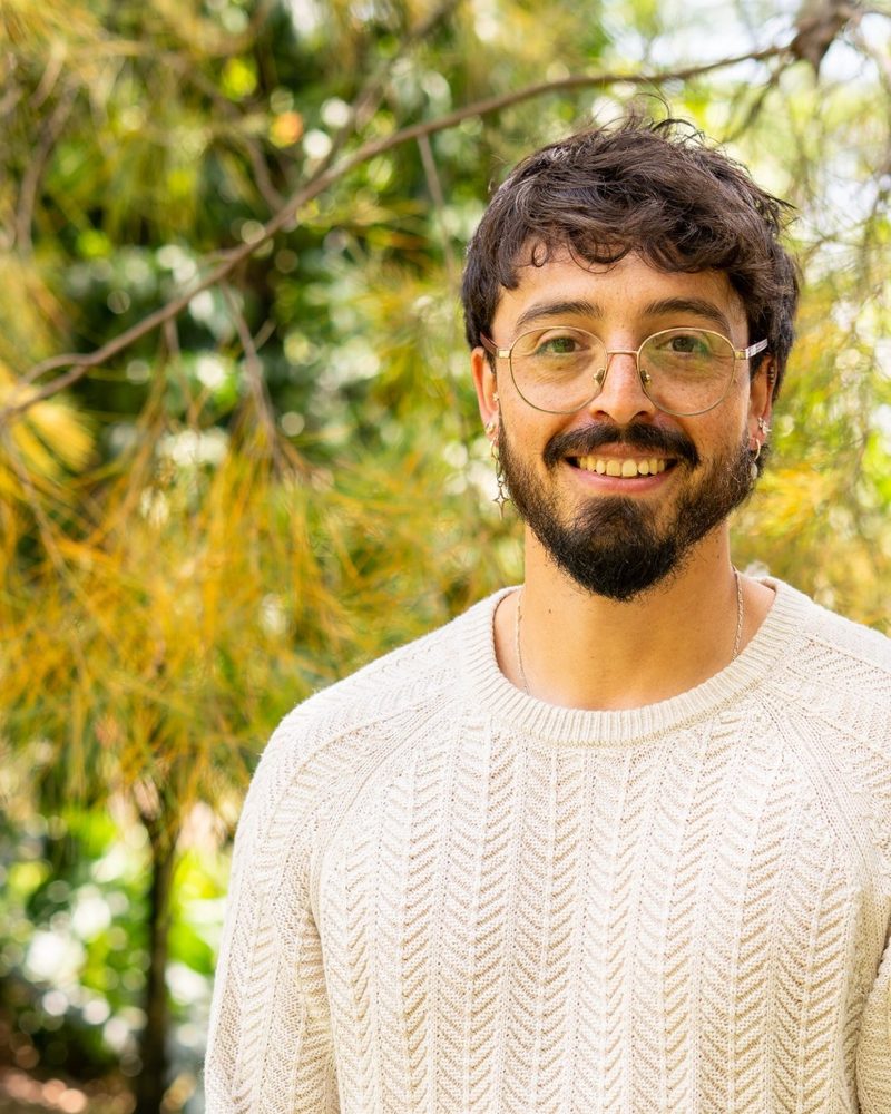 Retrato de Andrés Esteban Cárdenas, sonriente, con un suéter claro, entre ramas de pino.