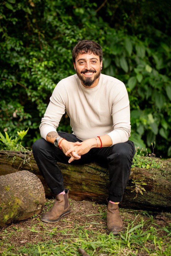 Andrés Esteban Cárdenas, sentado sobre un tronco en un jardín, sonríe a la cámara.