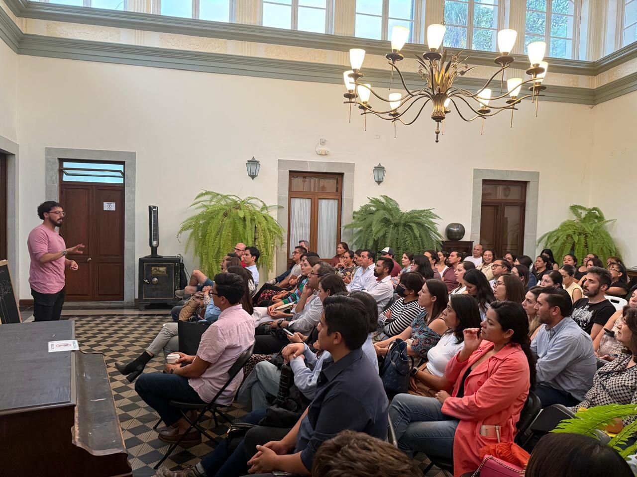 Andrés Esteban Cárdenas habla de pie ante una sala llena de personas adultas, en un salón con candelabro y ventanales.