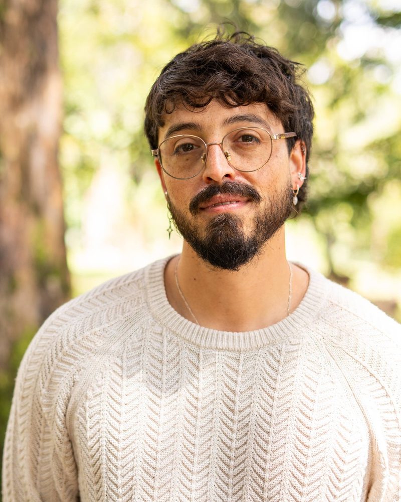 Retrato de Andrés Esteban Cárdenas en primer plano, con un suéter claro, junto a un árbol.