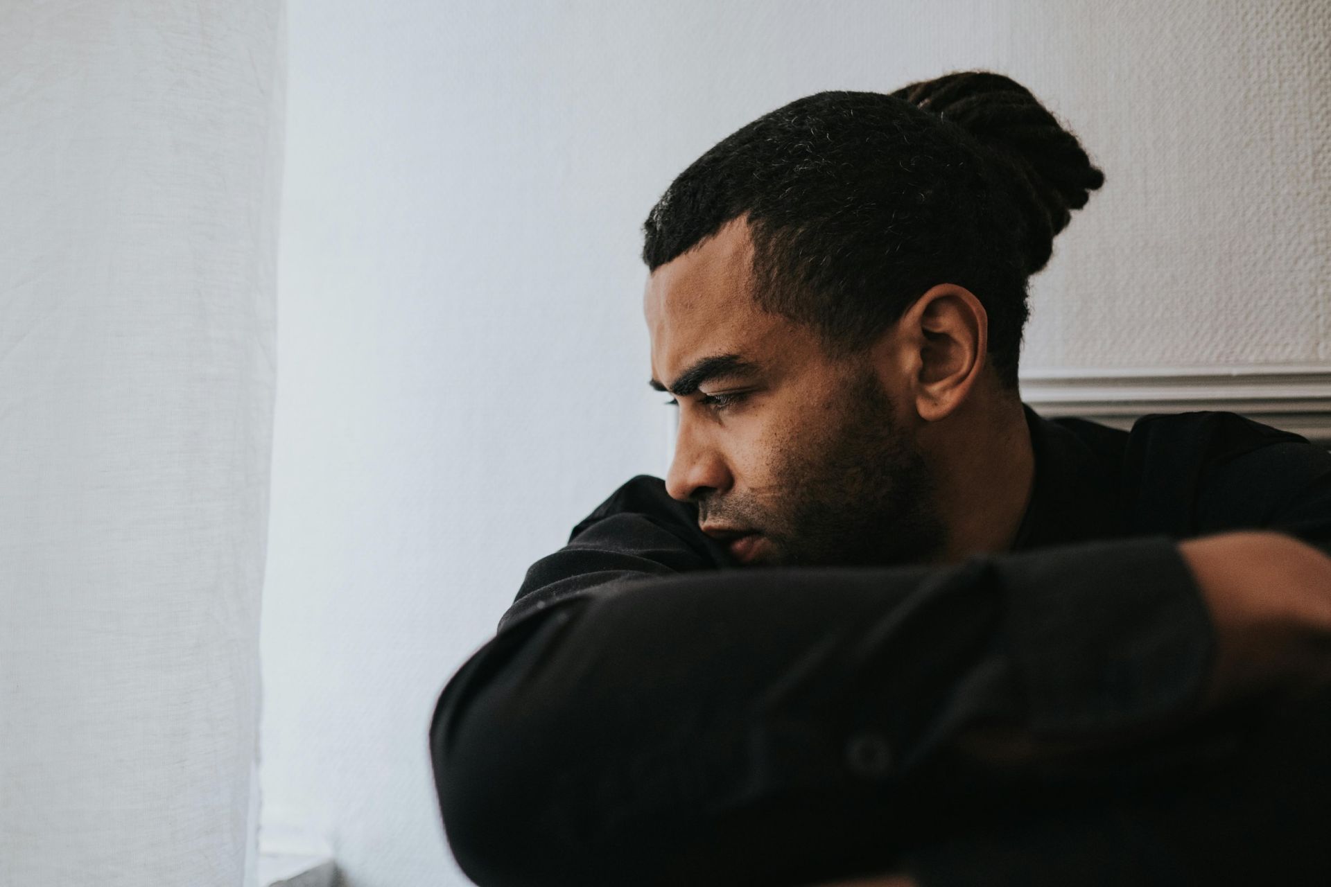A young Black man is curled up towards the crook of his elbow, staring despondently down at the ground
