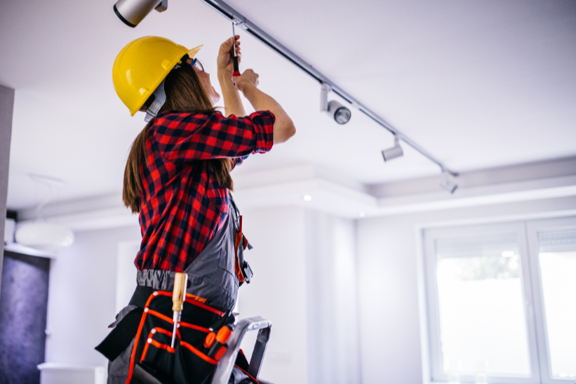 Professional electrician in safety gear installing LED track lighting in modern white interior, wearing yellow hard hat and plaid shirt RBQ 16