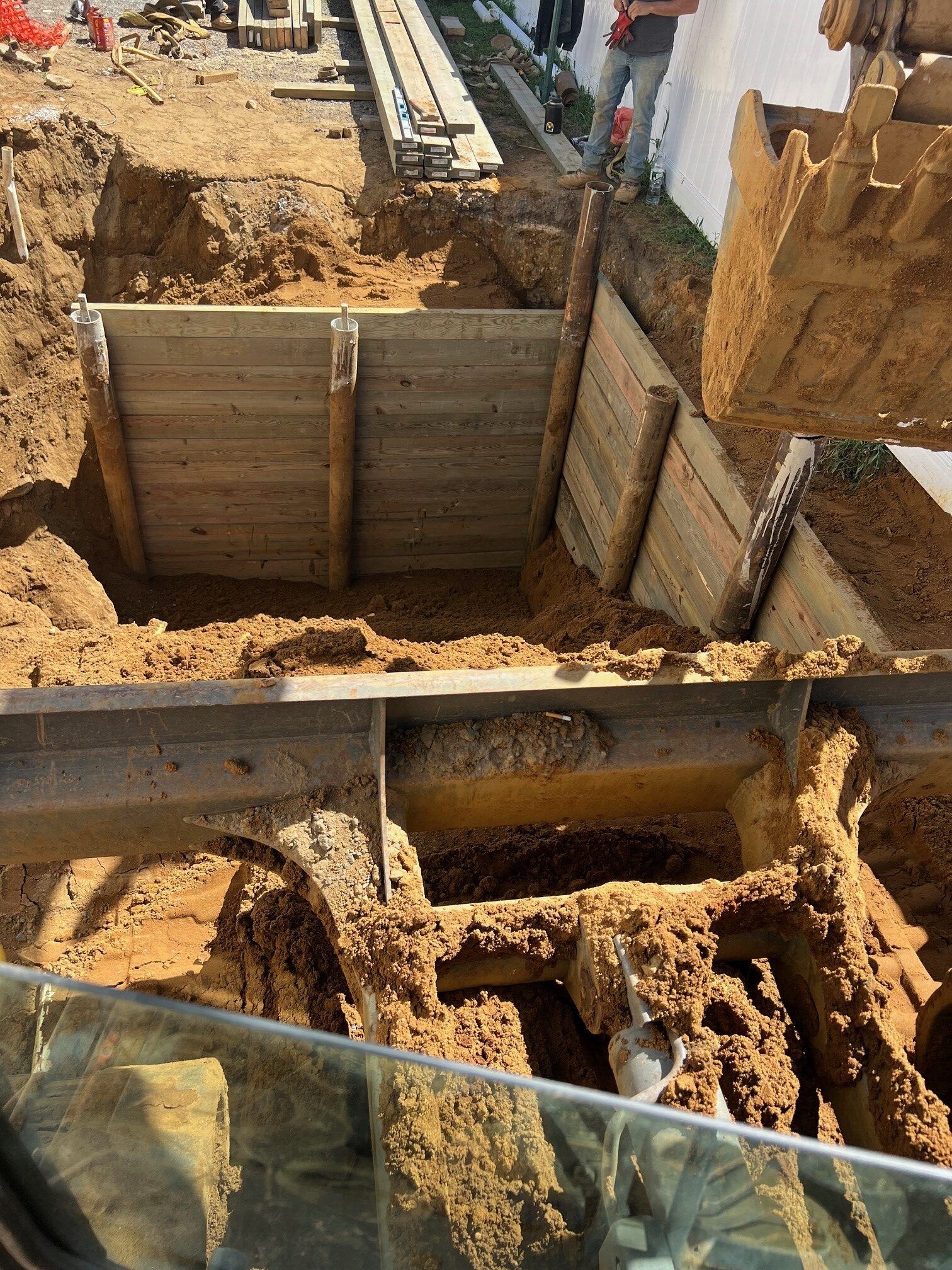 A construction site showing a deep excavation in soil with a newly installed wooden retaining wall. An excavator bucket is visible in the foreground, and a worker stands near the top edge in the background.
