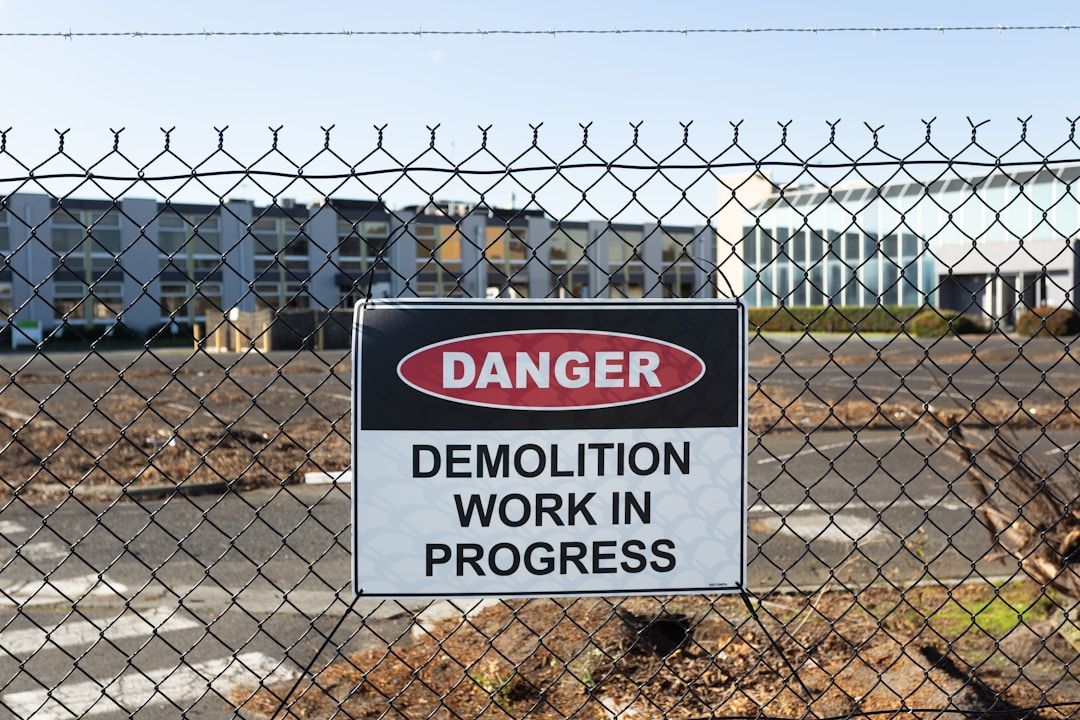 A DANGER sign on a chain-link fence that reads 'DEMOLITION WORK IN PROGRESS' in front of industrial-looking buildings.