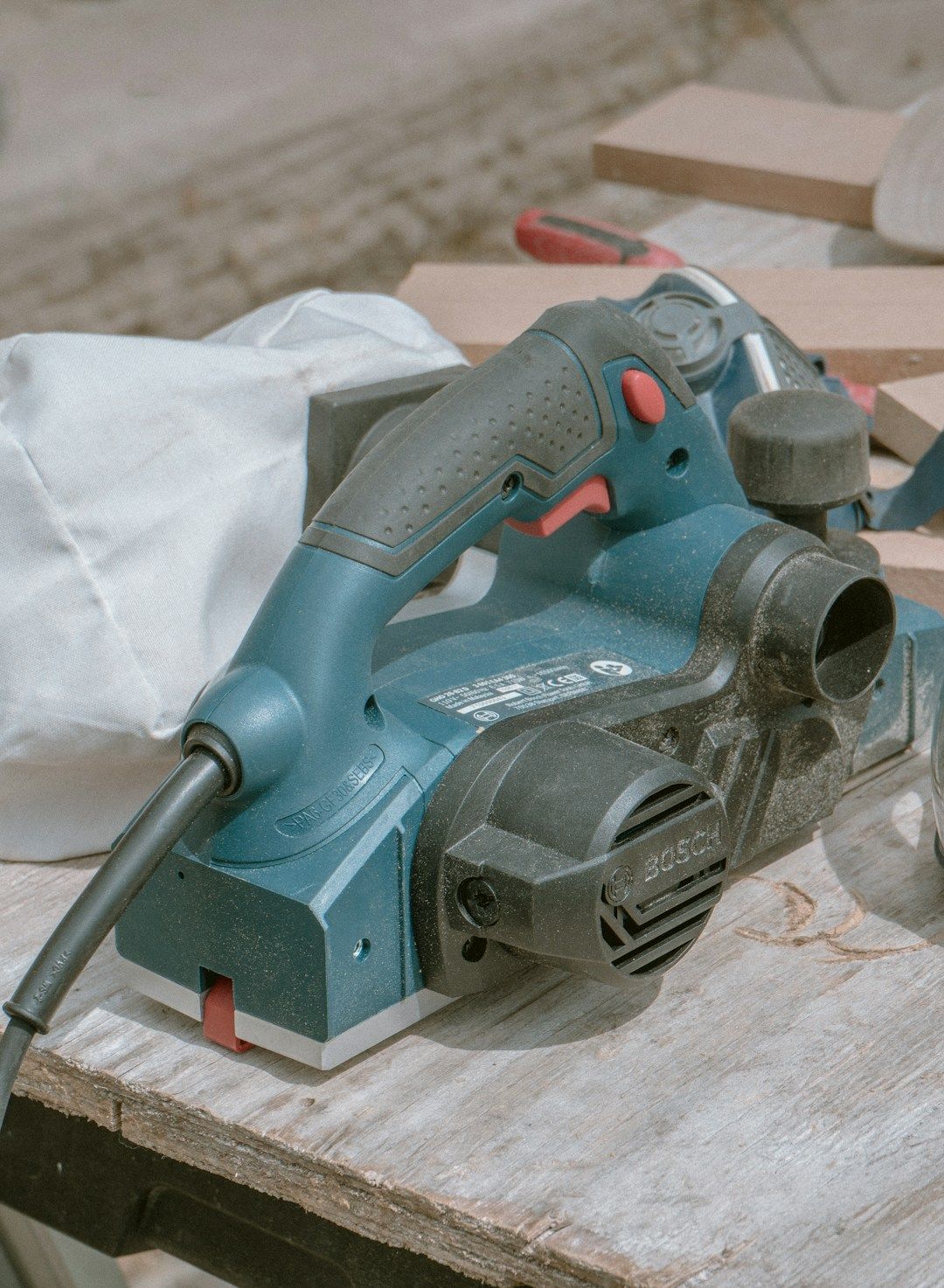 A blue and black Bosch electric hand planer is sitting on a wooden surface, with a white dust collection bag attached to its side and pieces of wood visible in the blurred background.