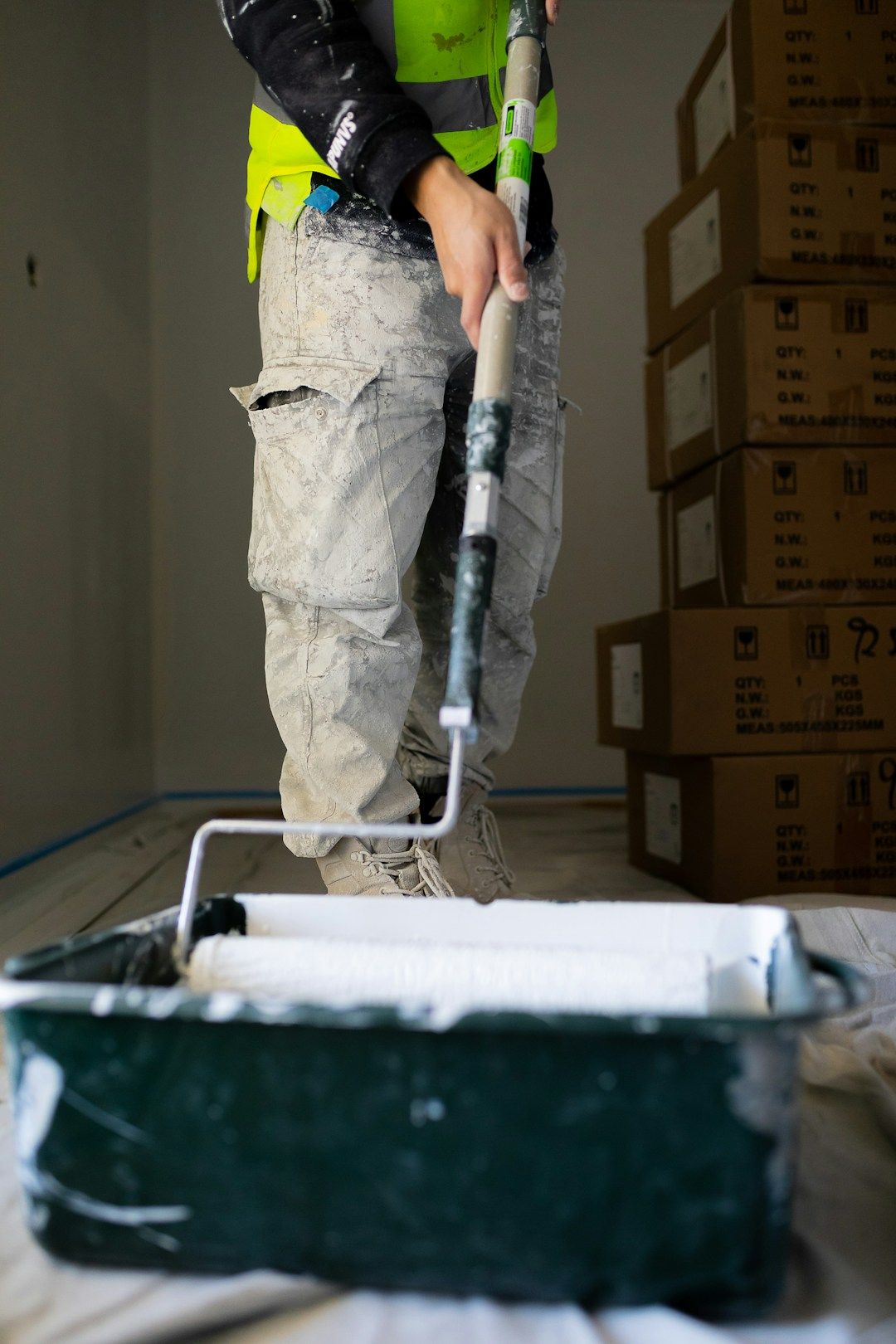 A worker in a high-visibility vest and camouflage pants uses a roller with an extension pole to apply white paint from a paint tray.