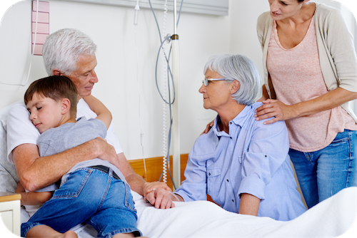 Mann im Krankenhaus wird von kleinem Jungen umarmt und zwei Frauen sitzen neben Krankenbett