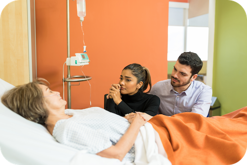 Ein junges Paar sitzt am Krankenbett einer schlafenden Frau
