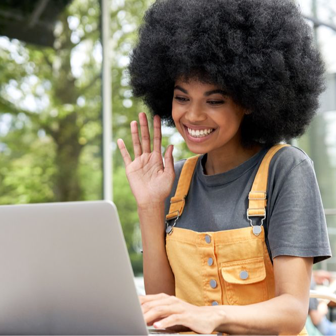  Hipster African American female student with Afro hair waving her hand while using a laptop computer for video conference call at an outdoor cafe table