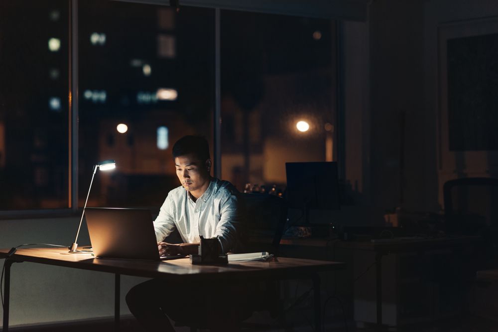 Young Asian businessman working on a laptop while sitting at his desk in a dark office at night with city lights in the background