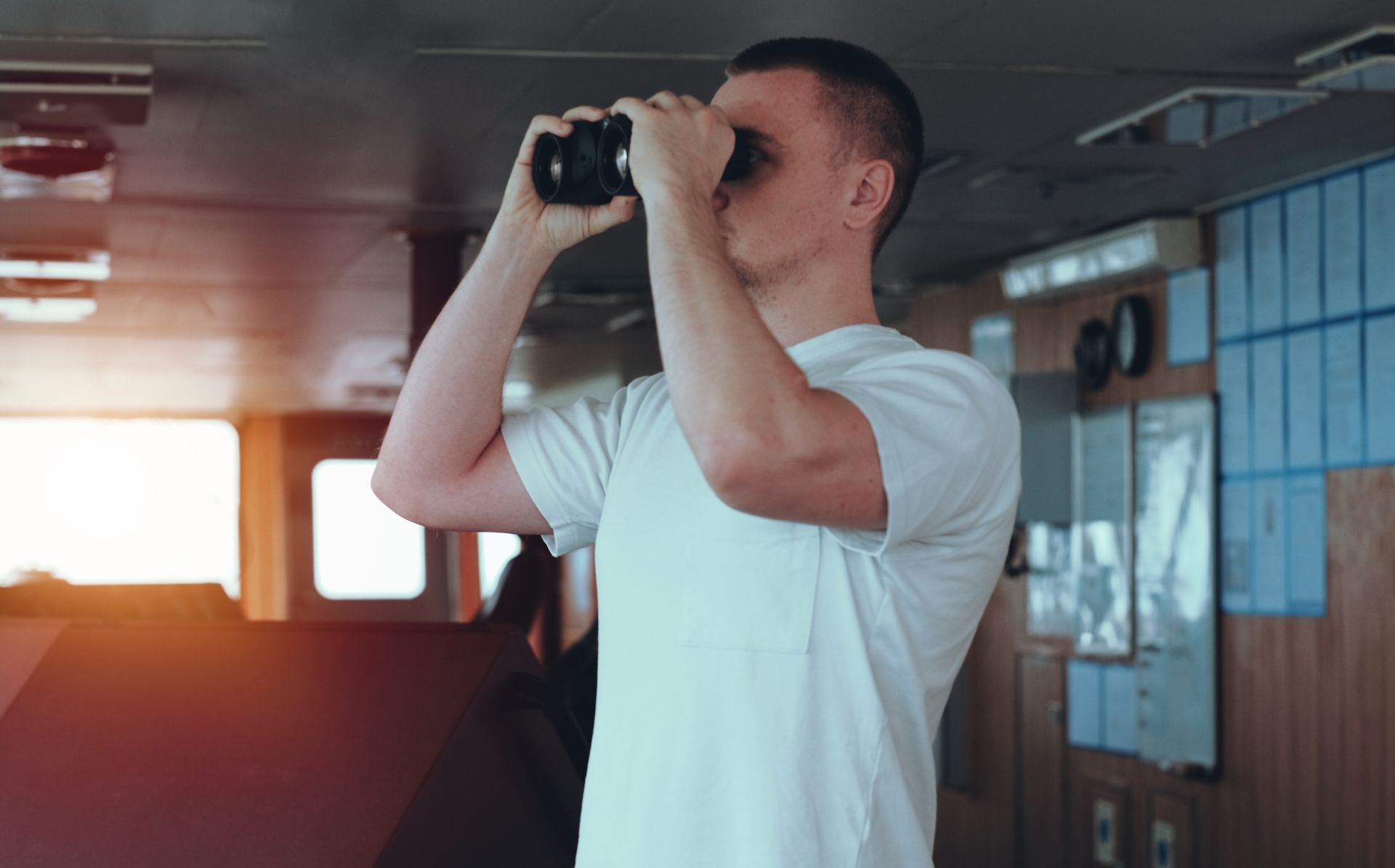 A young sailor in a white shirt using binoculars to look out from the ship's bridge, focusing on distant views for navigation or safety.
