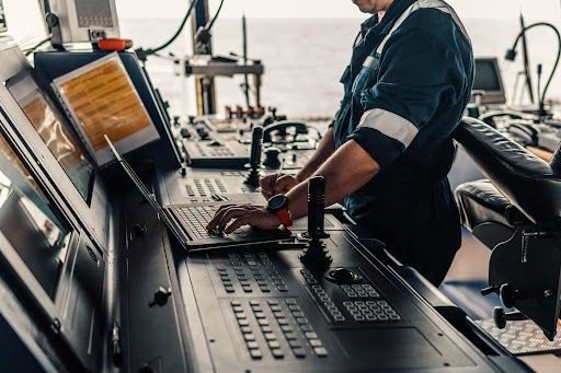 A maritime professional is operating complex navigation equipment and using a laptop on the bridge of a ship, highlighting modern seafaring technology.