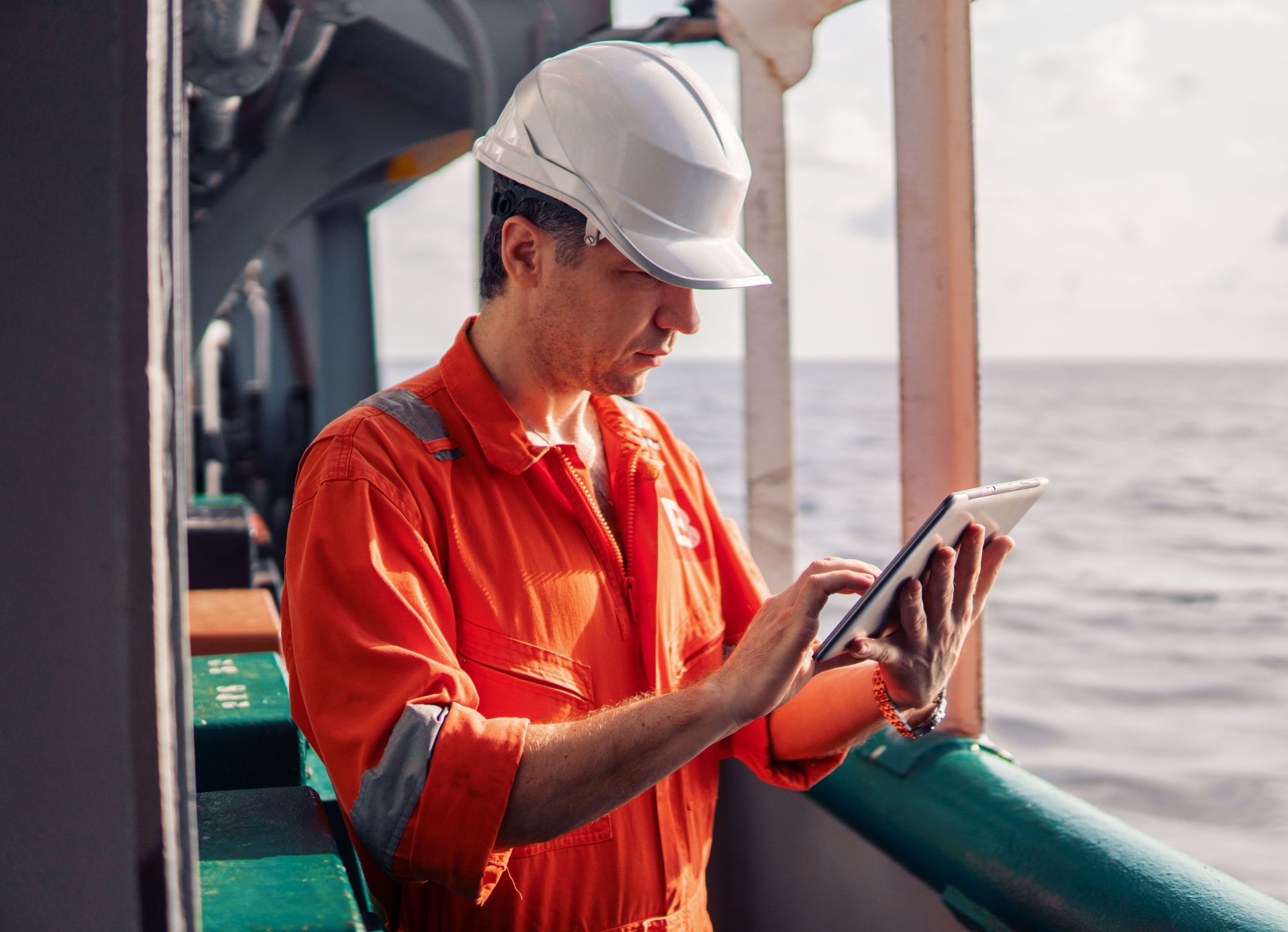 Marine engineer in orange coveralls using a tablet on the deck of a cargo ship.