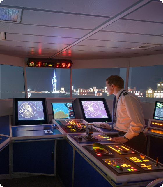 A seafarer operating navigational equipment in a ship's bridge simulator with multiple radar screens, control panels, and a view of a simulated coastal cityscape at night