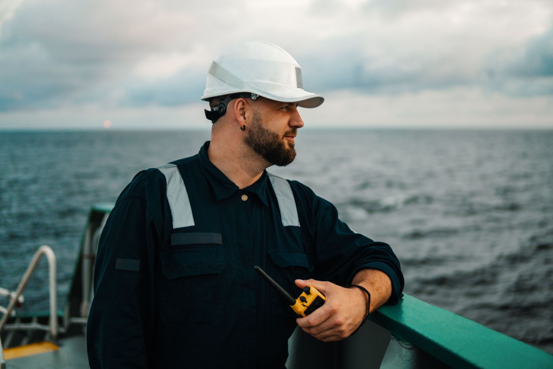 Maritime professional with a hard hat and walkie-talkie looking out at sea, standing by the ship's railing at sunset.
