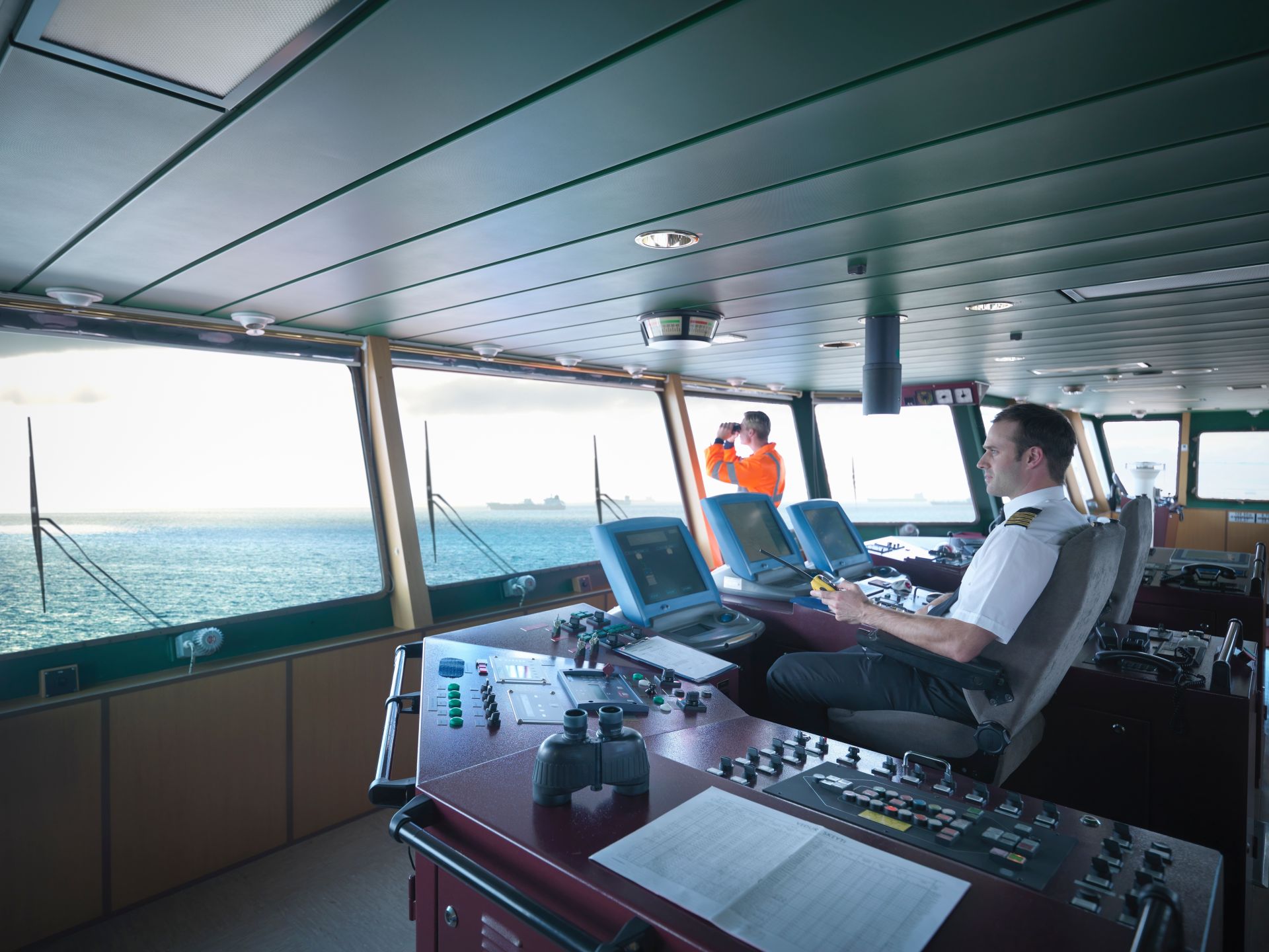 Inside the bridge of a ship with a captain operating navigational equipment while a crew member looks out to sea with binoculars, indicating active maritime navigation.