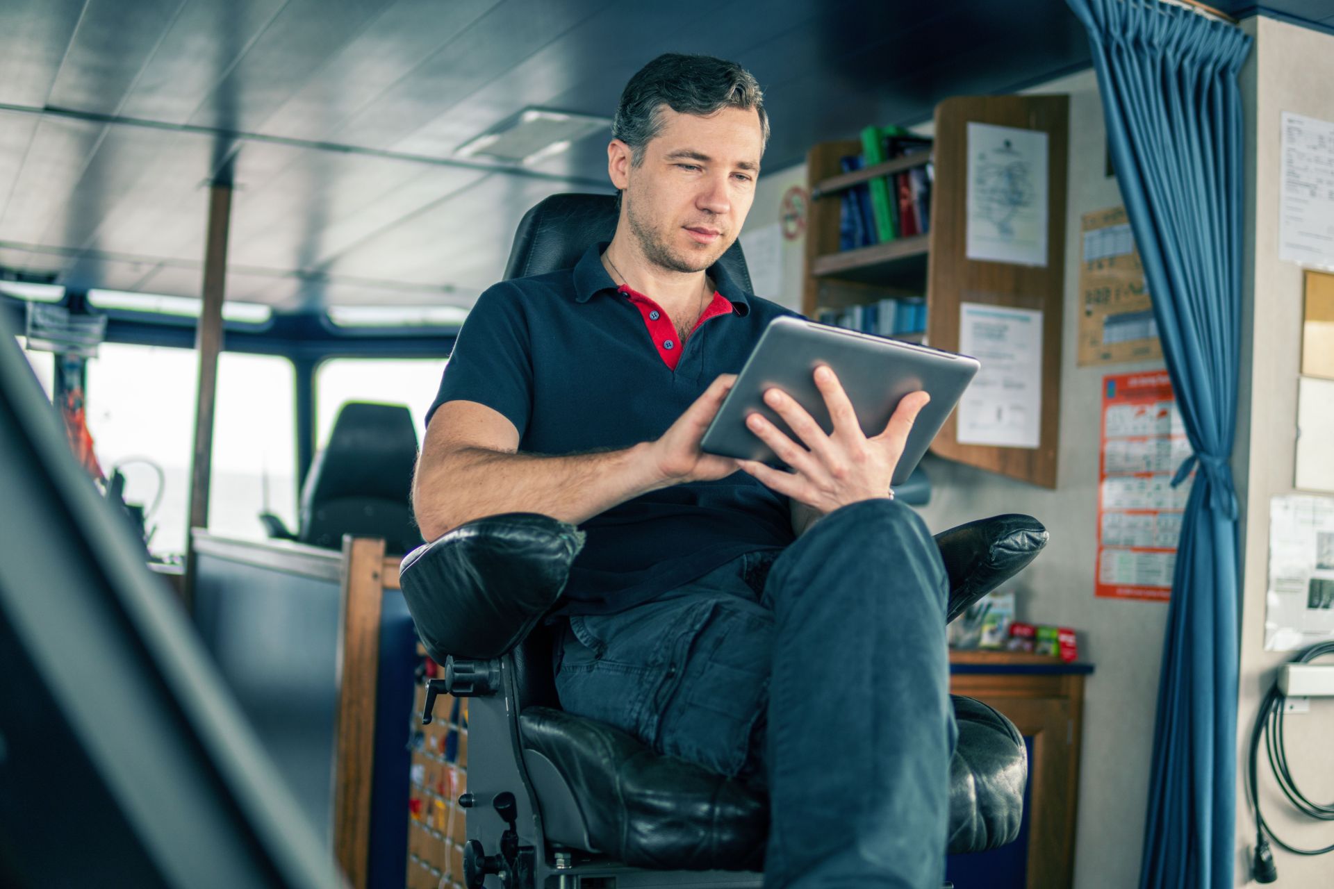 A seafarer in casual attire relaxes in a chair on the ship's bridge, deeply engrossed in reading or working on a tablet device.