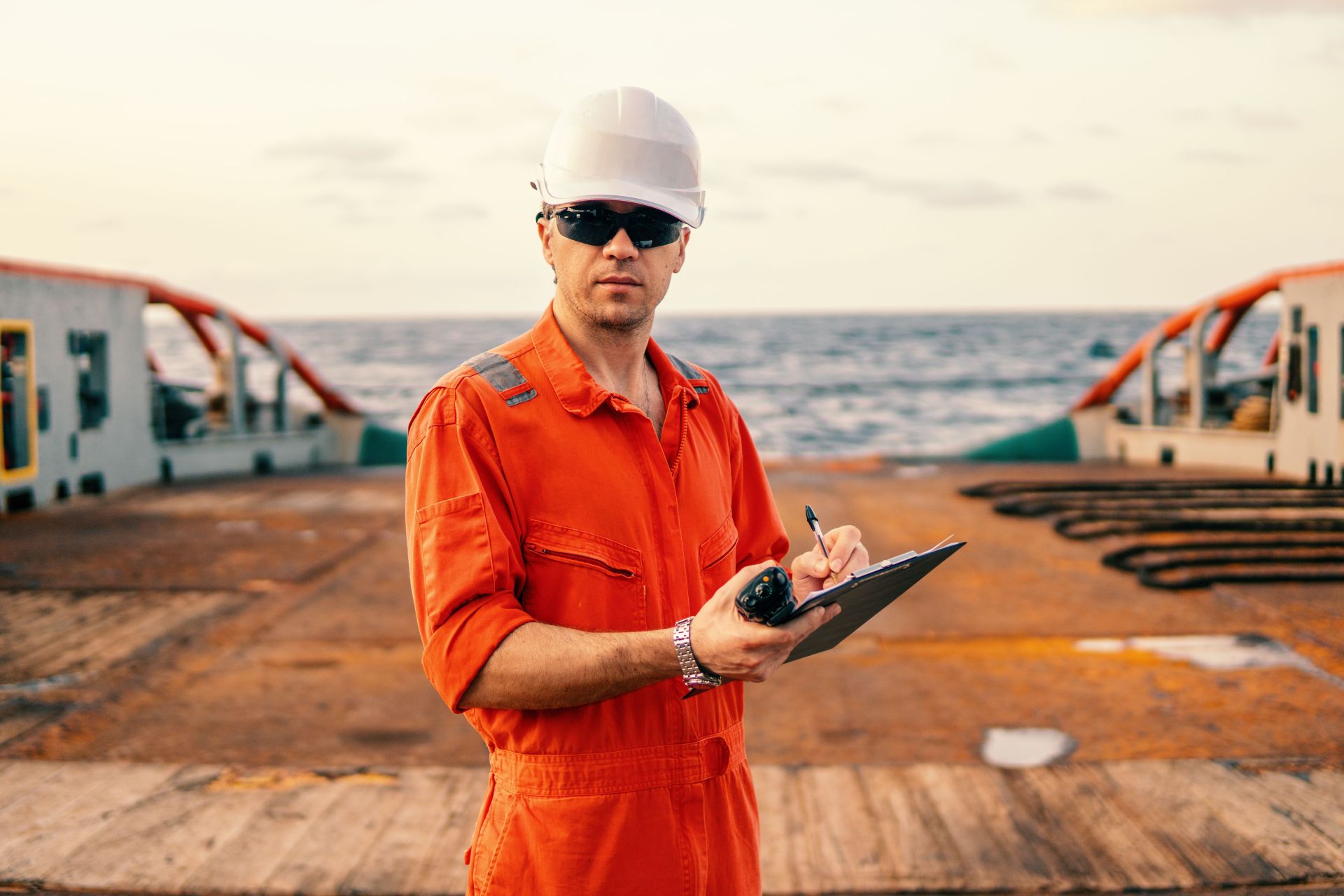 Marine engineer in orange coveralls and white hard hat holding a clipboard and a handheld VHF radio on the deck of a ship, with the ocean in the background.