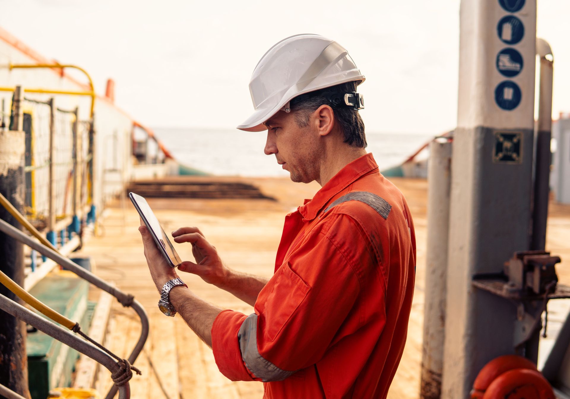 A maritime worker in orange coveralls and a white safety helmet attentively uses a tablet on the deck of a vessel, possibly for inspection or operational tasks.