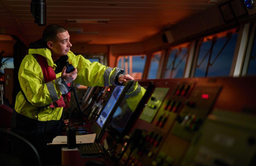 A maritime professional is operating complex navigation equipment and using a laptop on the bridge of a ship, highlighting modern seafaring technology.