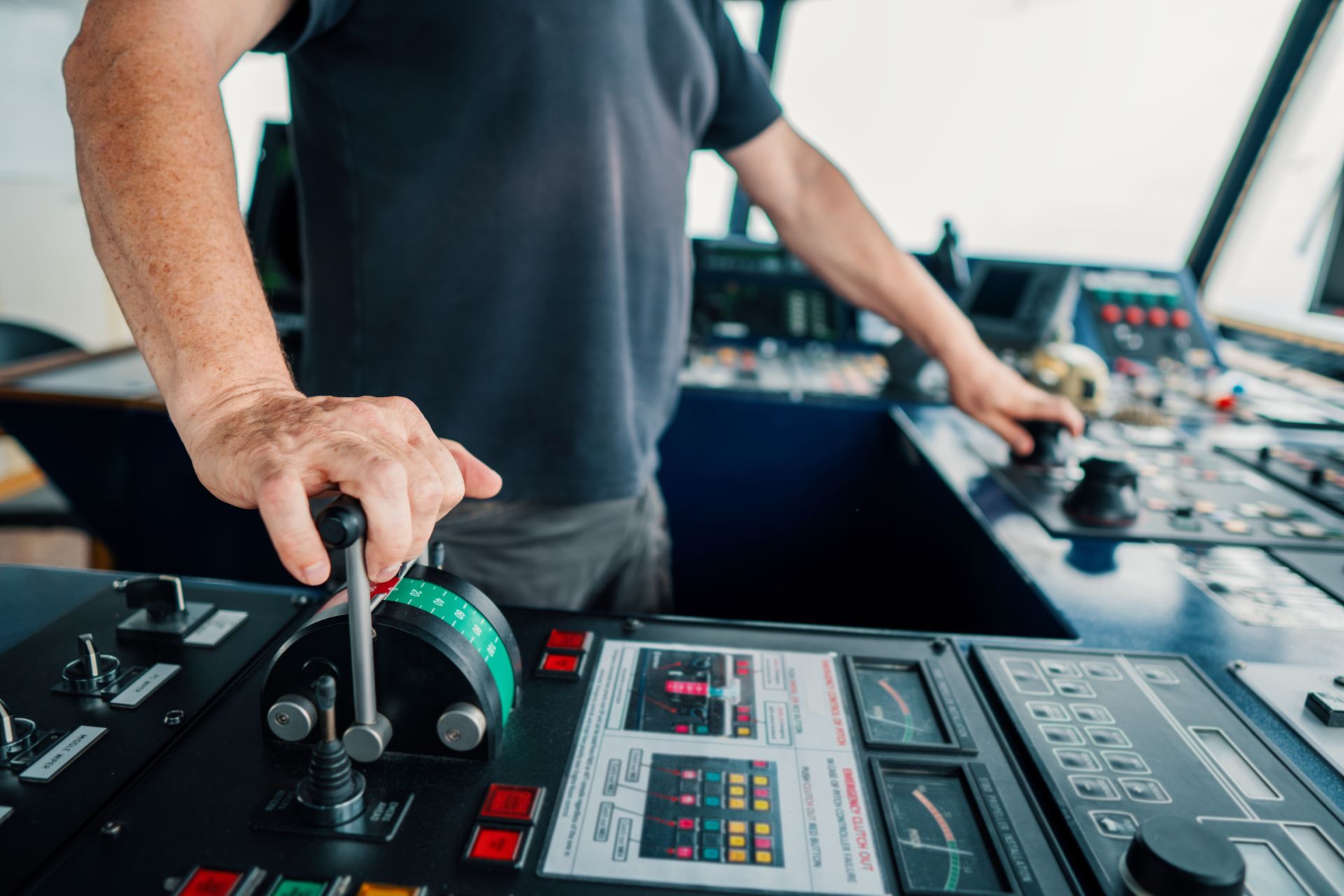 A close-up of a mariner's hands operating ship control levers on a vessel's bridge, with navigation panels and monitors in the background, indicating active maritime operation.