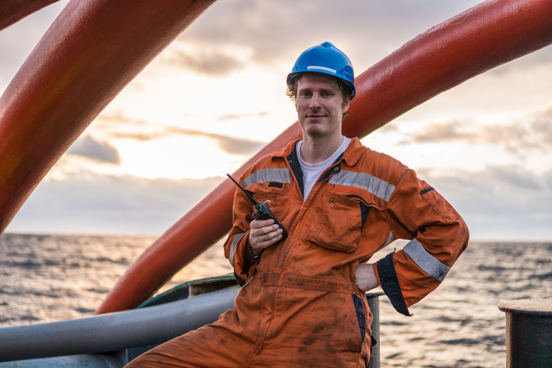 Confident maritime crew member in orange overalls and blue helmet holding a walkie-talkie on the ship deck at dawn.