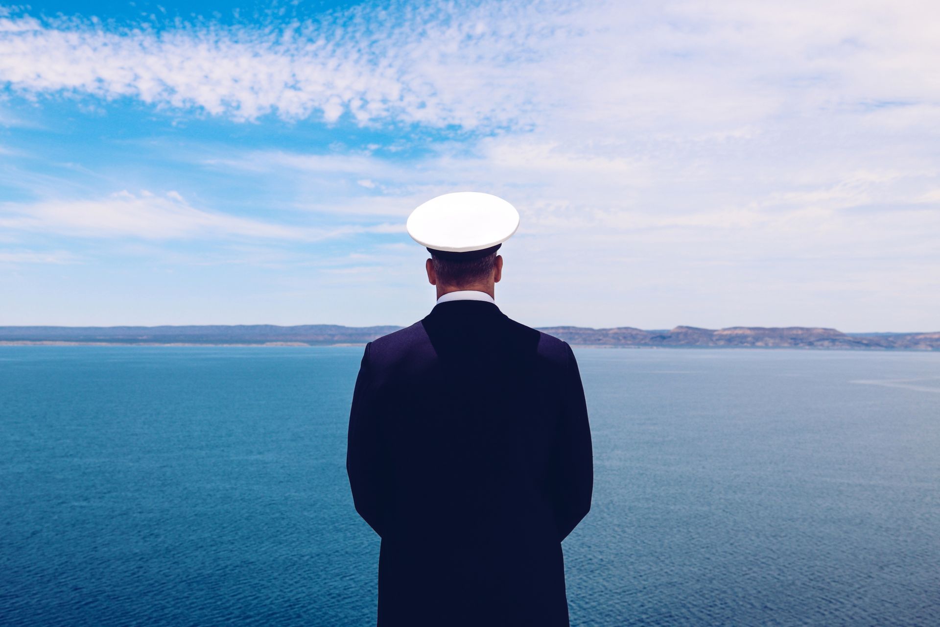 Rear view of a ship's officer in a navy blue uniform and white cap looking out over the calm blue sea, symbolizing leadership and vigilance at sea.